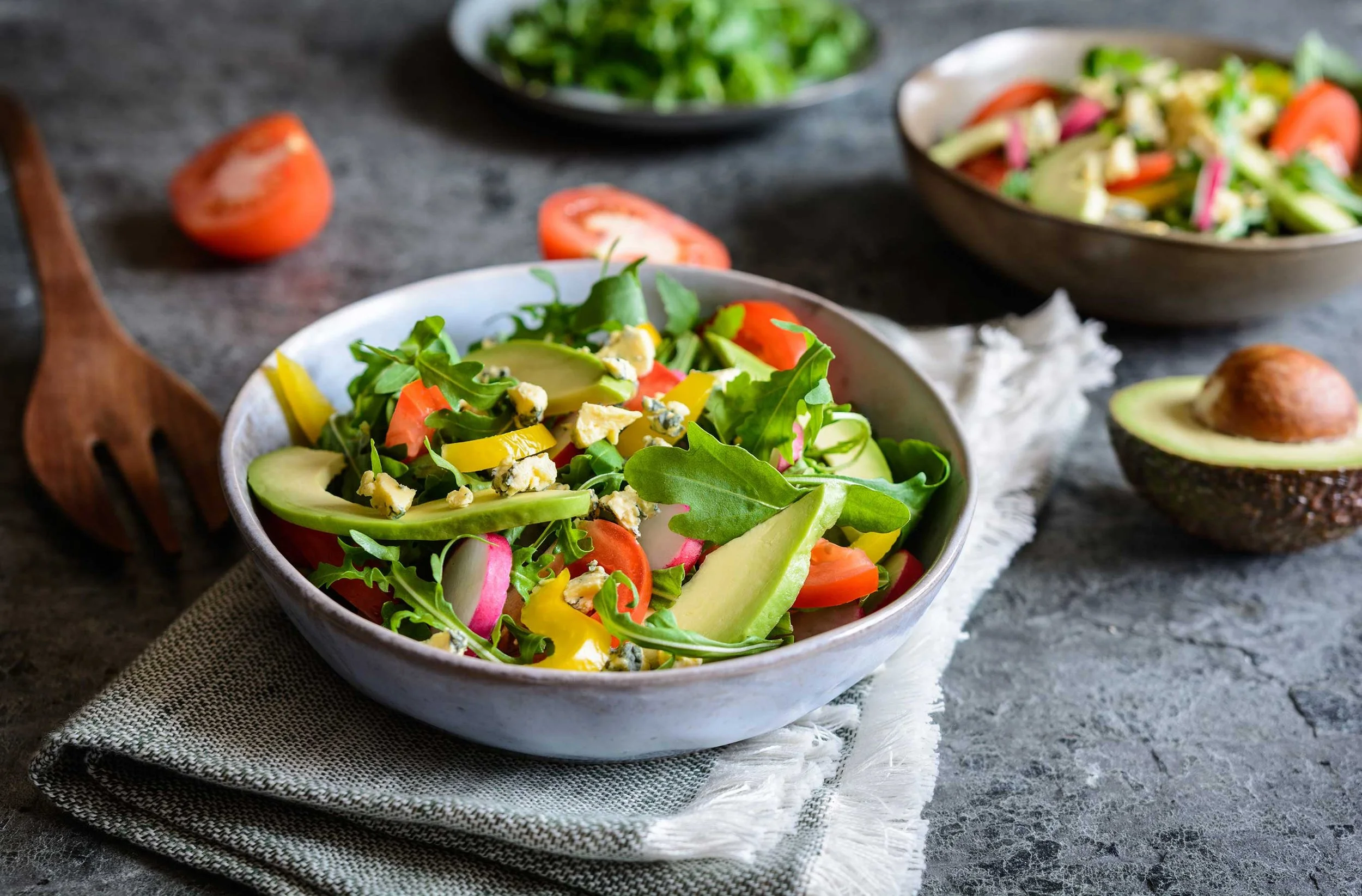 A bowl of fresh mixed vegetable salad on a white napkin, with sliced tomatoes and avocado, on a dark textured surface.