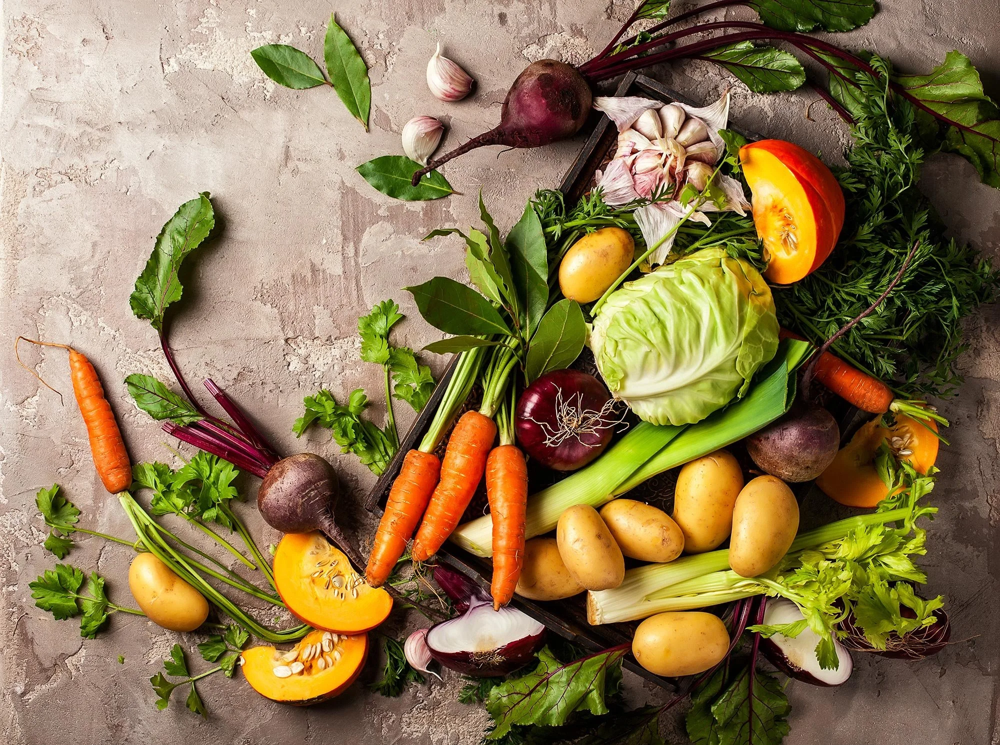 A top-down view of various fresh vegetables including carrots, potatoes, beetroot, garlic, bell peppers, lettuce, and herbs scattered on a textured surface.