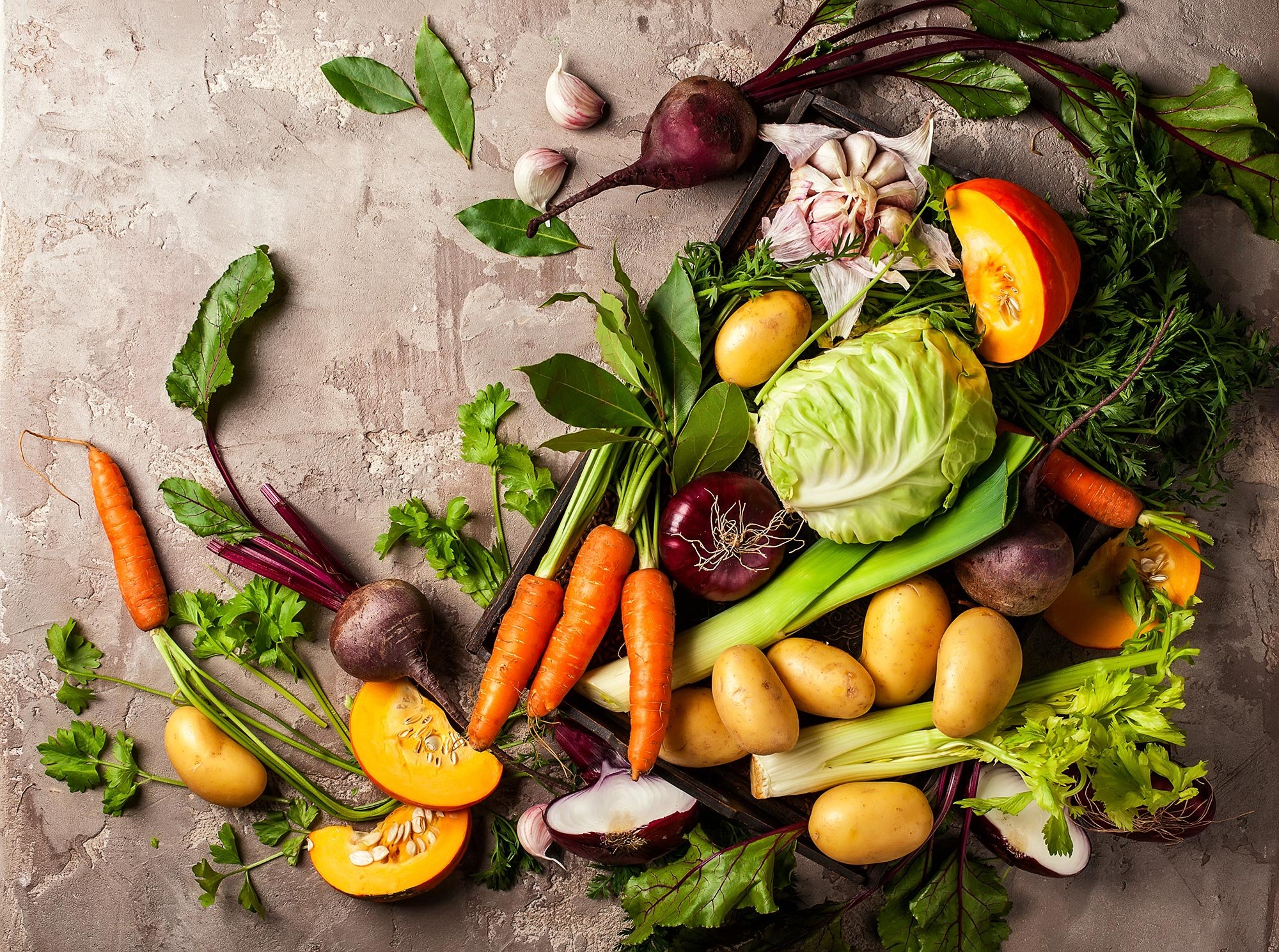 A variety of fresh vegetables including carrots, potatoes, beets, garlic, cabbage, and bell peppers, arranged on a textured surface.