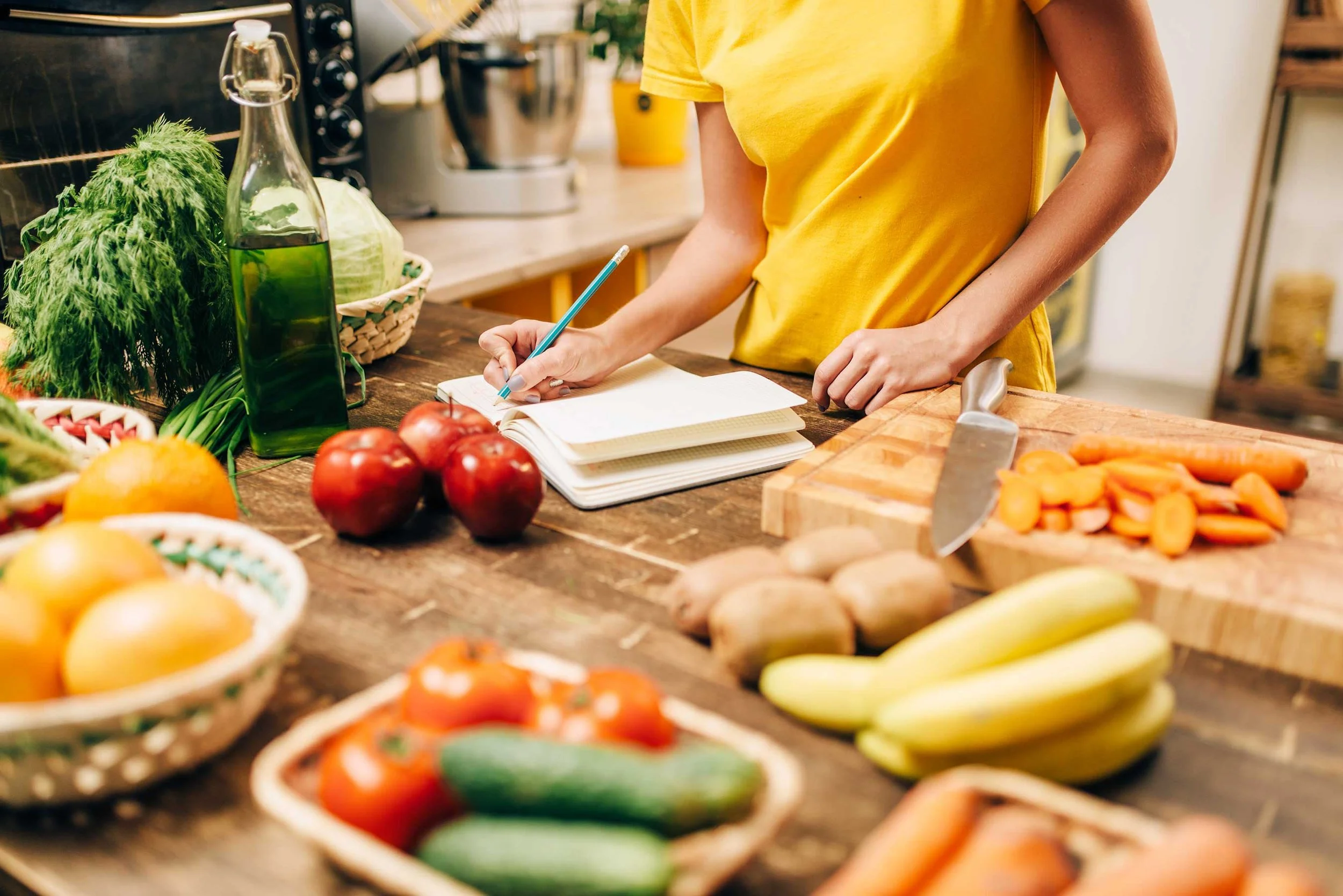 Person in yellow shirt writing in a notebook on a kitchen counter surrounded by fresh vegetables including tomatoes, cucumbers, carrots, bananas, potatoes, and a bottle of oil.