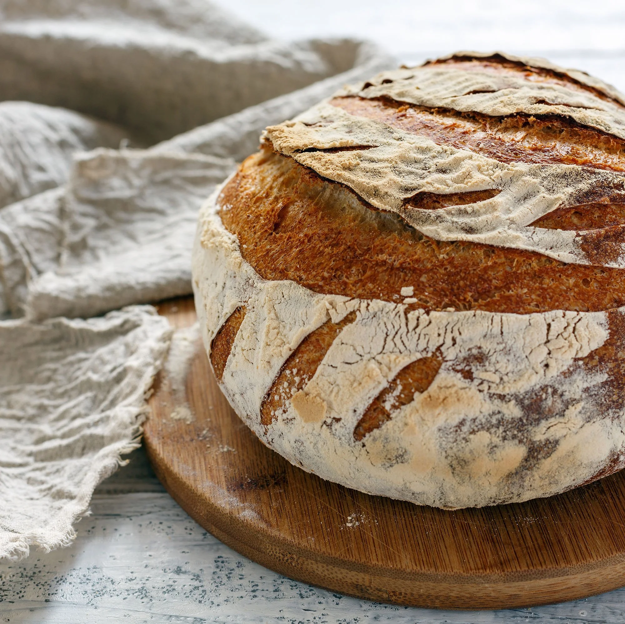 Freshly baked loaf of bread with a golden crust resting on a wooden cutting board, sprinkled with flour, with a rustic cloth in the background.