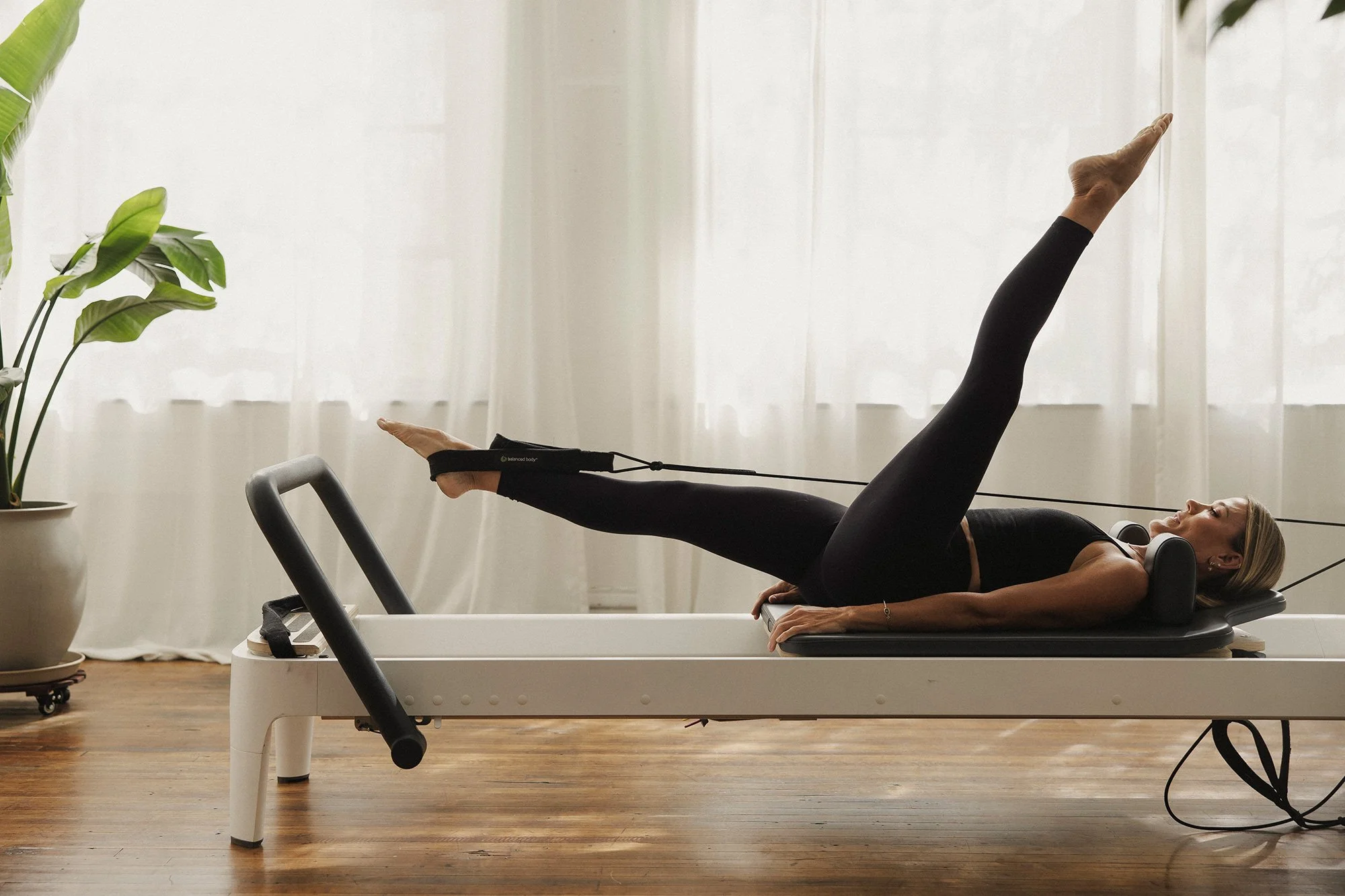 Woman exercising on a Pilates reformer machine in a bright room with large windows and sheer curtains, with a potted plant nearby.