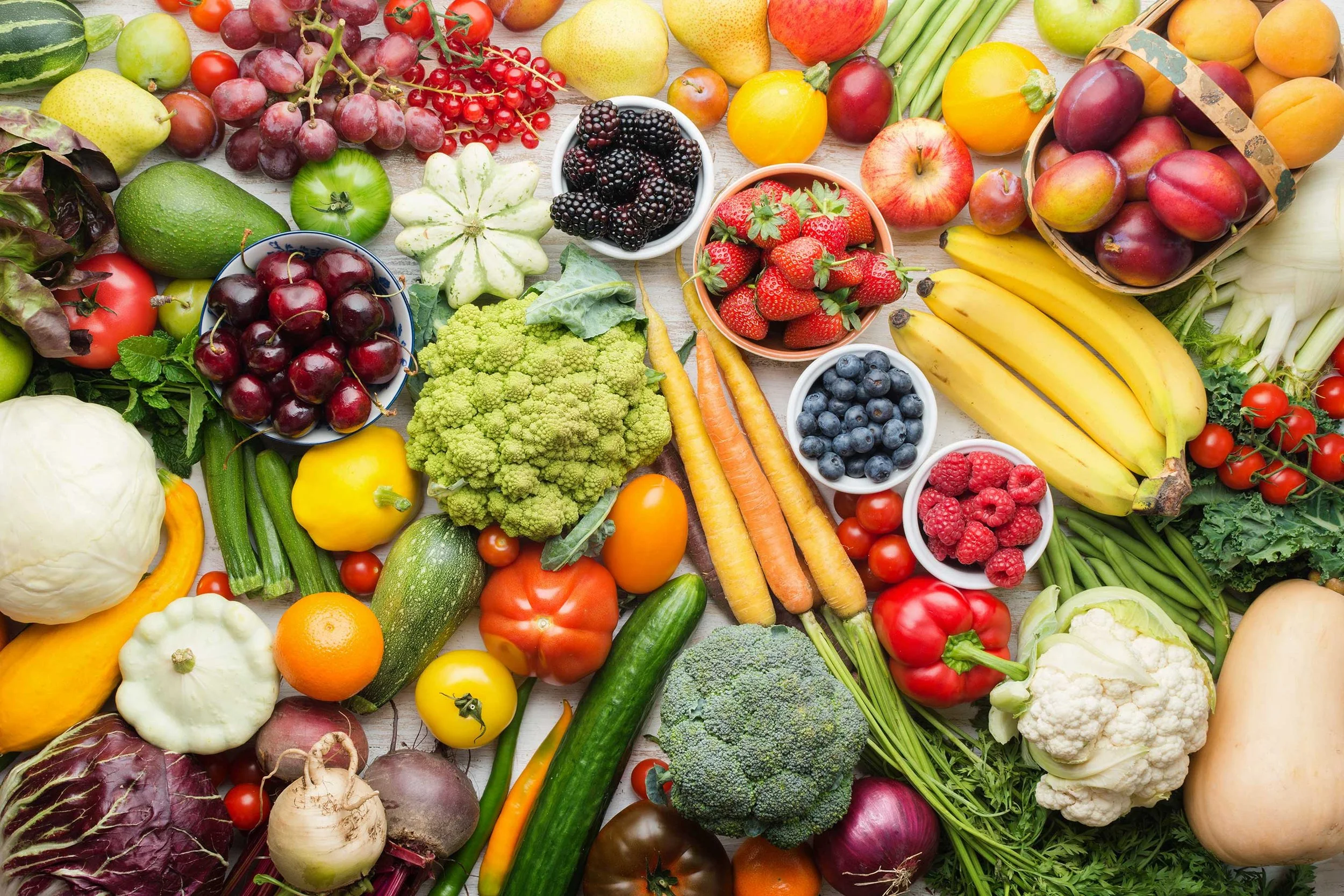 Assorted fresh fruits and vegetables spread on a wooden surface, including strawberries, blueberries, blackberries, cherries, grapes, bananas, broccoli, cauliflower, carrots, cucumbers, tomatoes, apples, and lettuce.