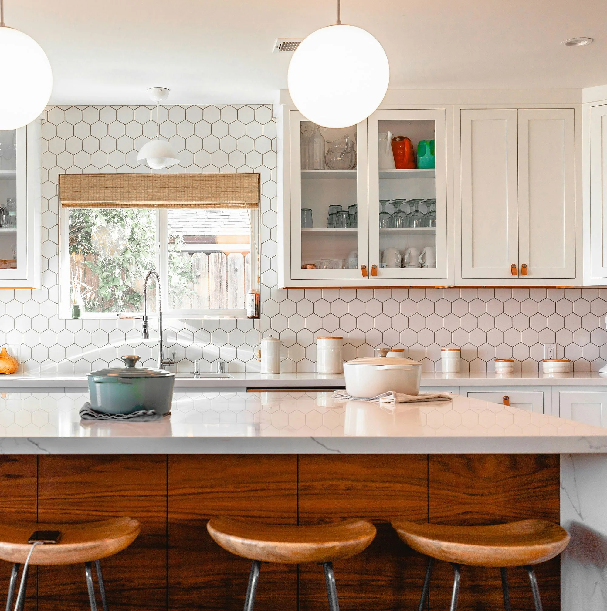 Bright kitchen with white cabinets, hexagon tiled backsplash, window with bamboo shade, and wooden stools at a white countertop. Hanging pendant and ceiling lights are visible.