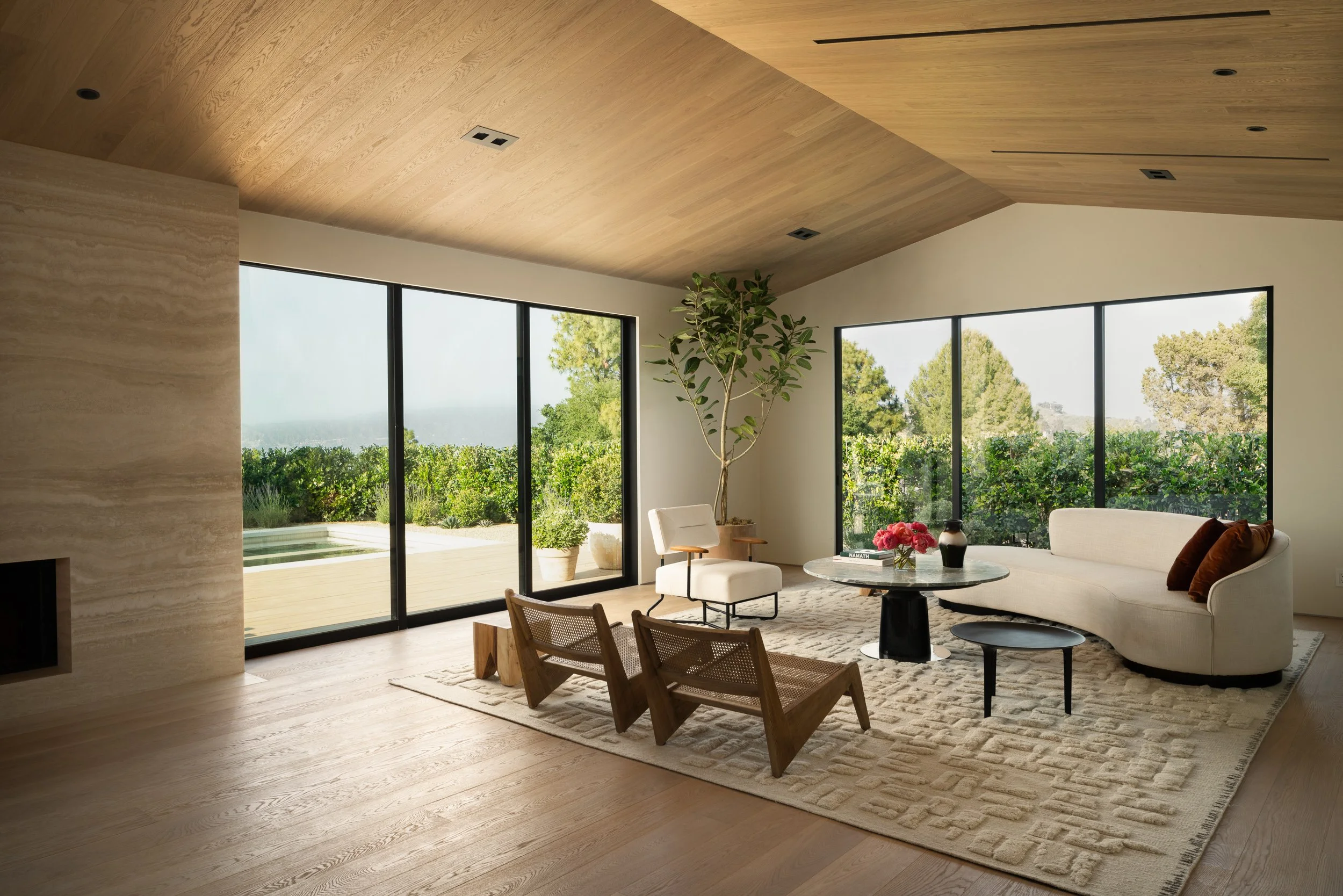 Modern living room with large glass doors, light wood flooring and ceiling, a white sofa, a round coffee table with pink flowers, and a tall potted plant outdoors