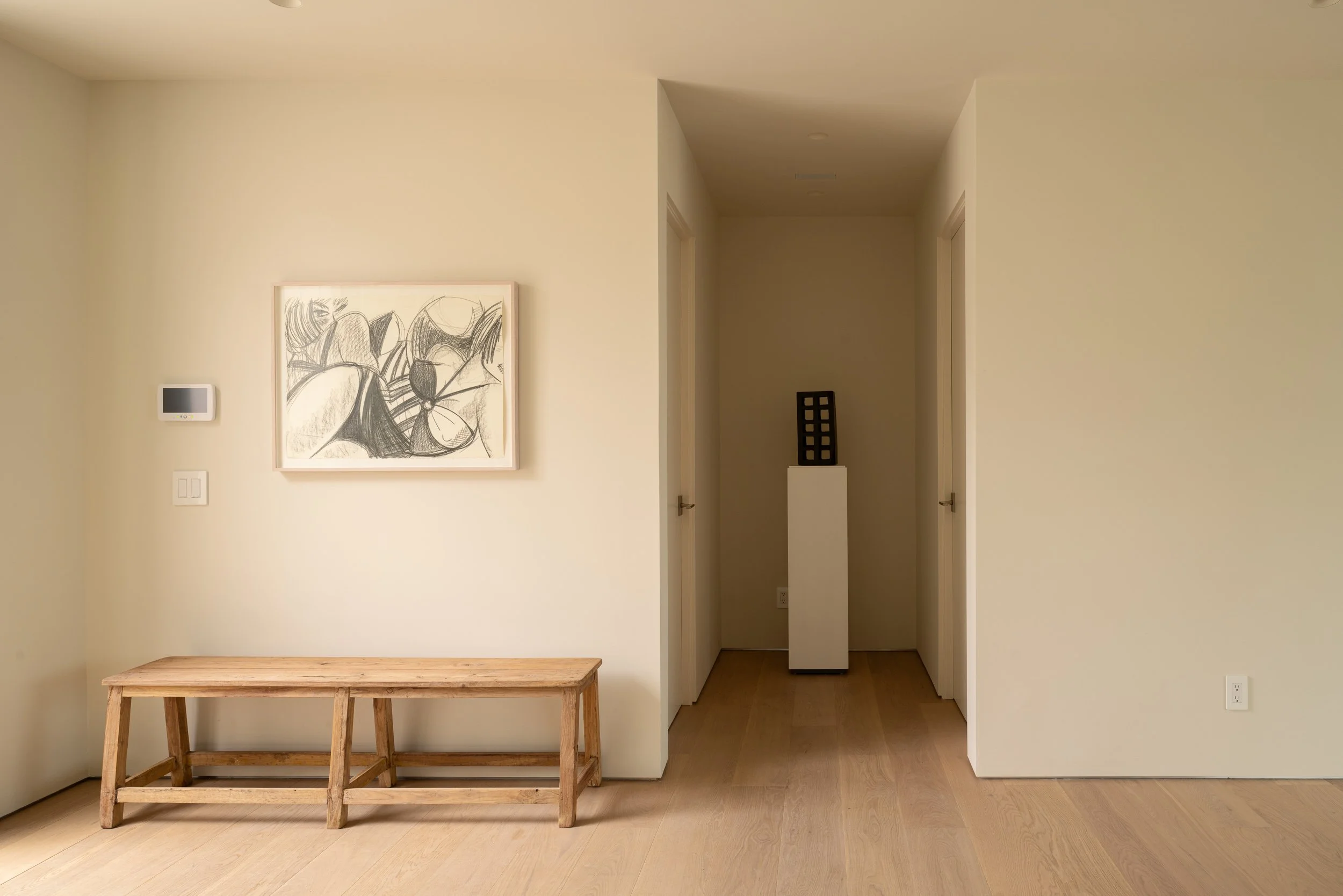 A minimalist room with off-white walls and light wooden flooring. There is a simple wooden bench in the foreground, a framed abstract black and white sketch hanging on the wall, and a small stop control panel beside it. The background features two white-doorways leading to a hallway with a small black decorative object on a white pedestal. The space is well-lit with no windows visible.