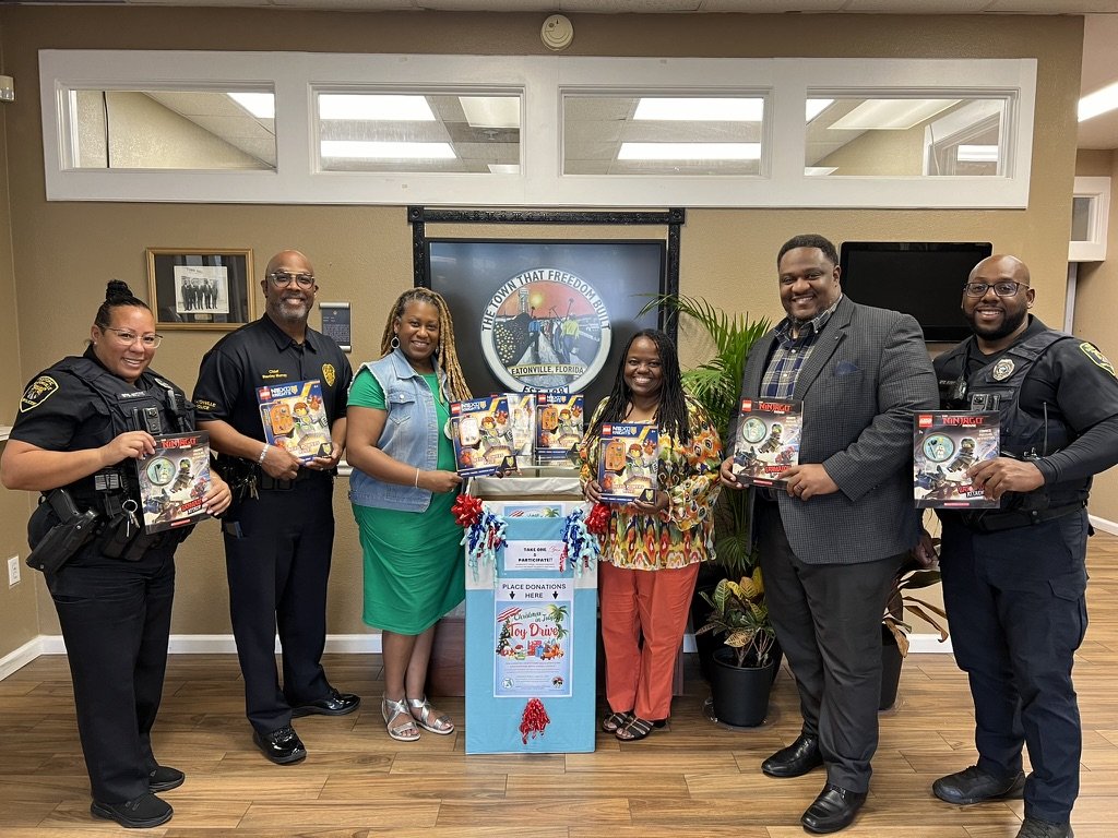 Group of six people, including police officers and civilians, standing in an office holding toy gift sets for a toy drive. A donation box and a sign for the toy drive are in front of them.