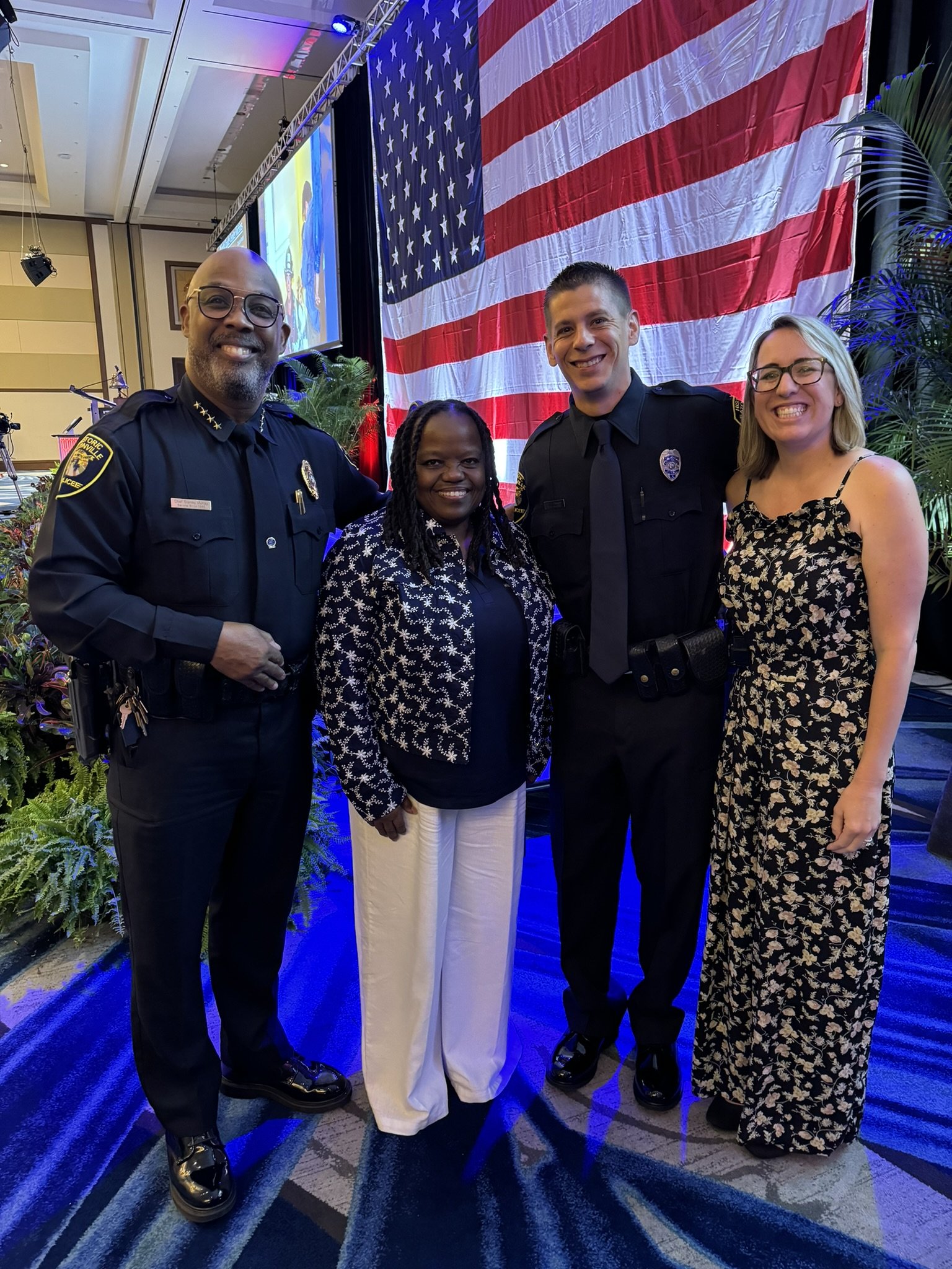 Four people standing together in front of a large American flag, smiling at the camera, at an indoor event with a stage and plants in the background.
