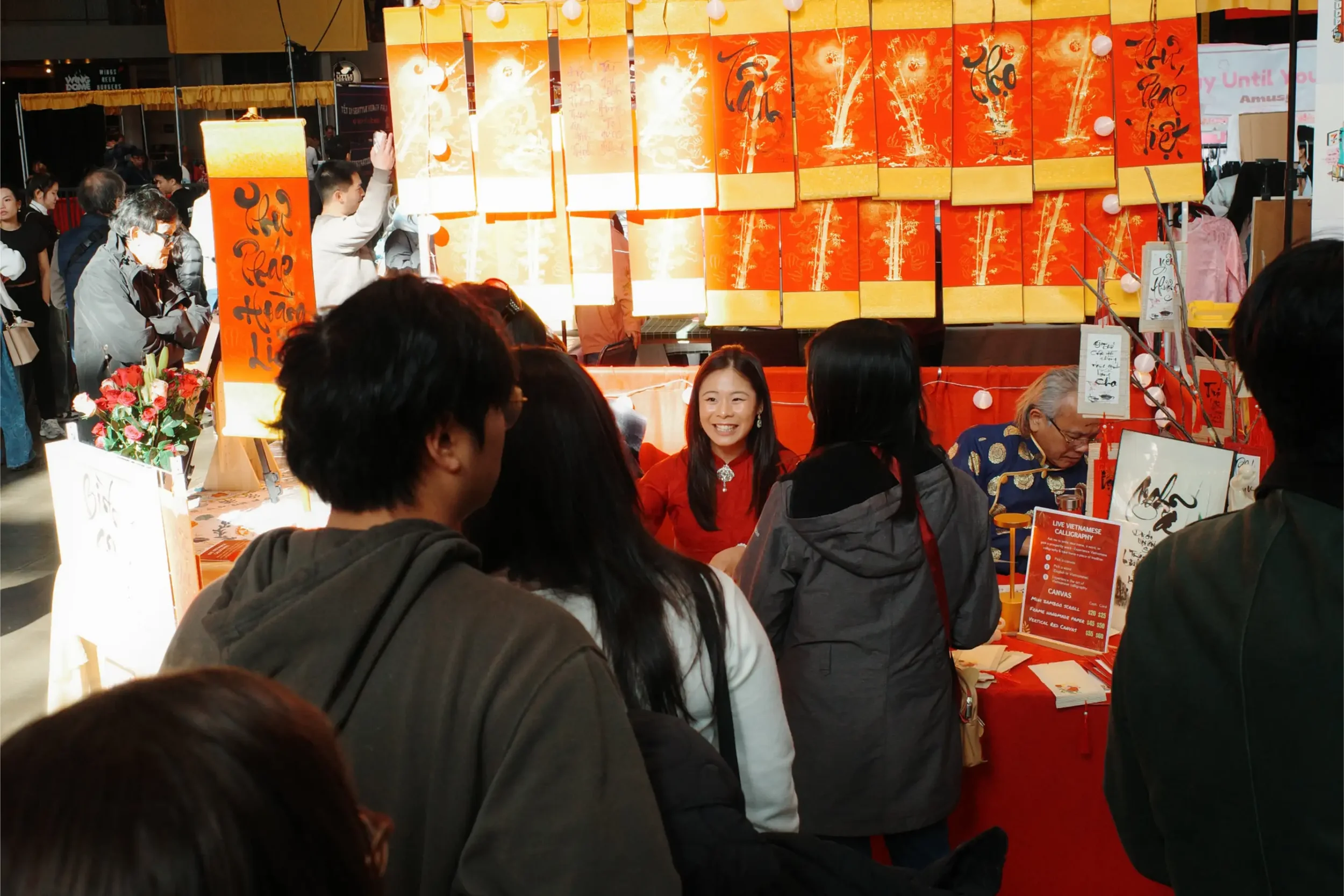 A crowd gathering at Linh Nguyen's live Vietnamese calligraphy booth at Tết Seattle 2025, with large orange and gold calligraphy scrolls displayed in the background.