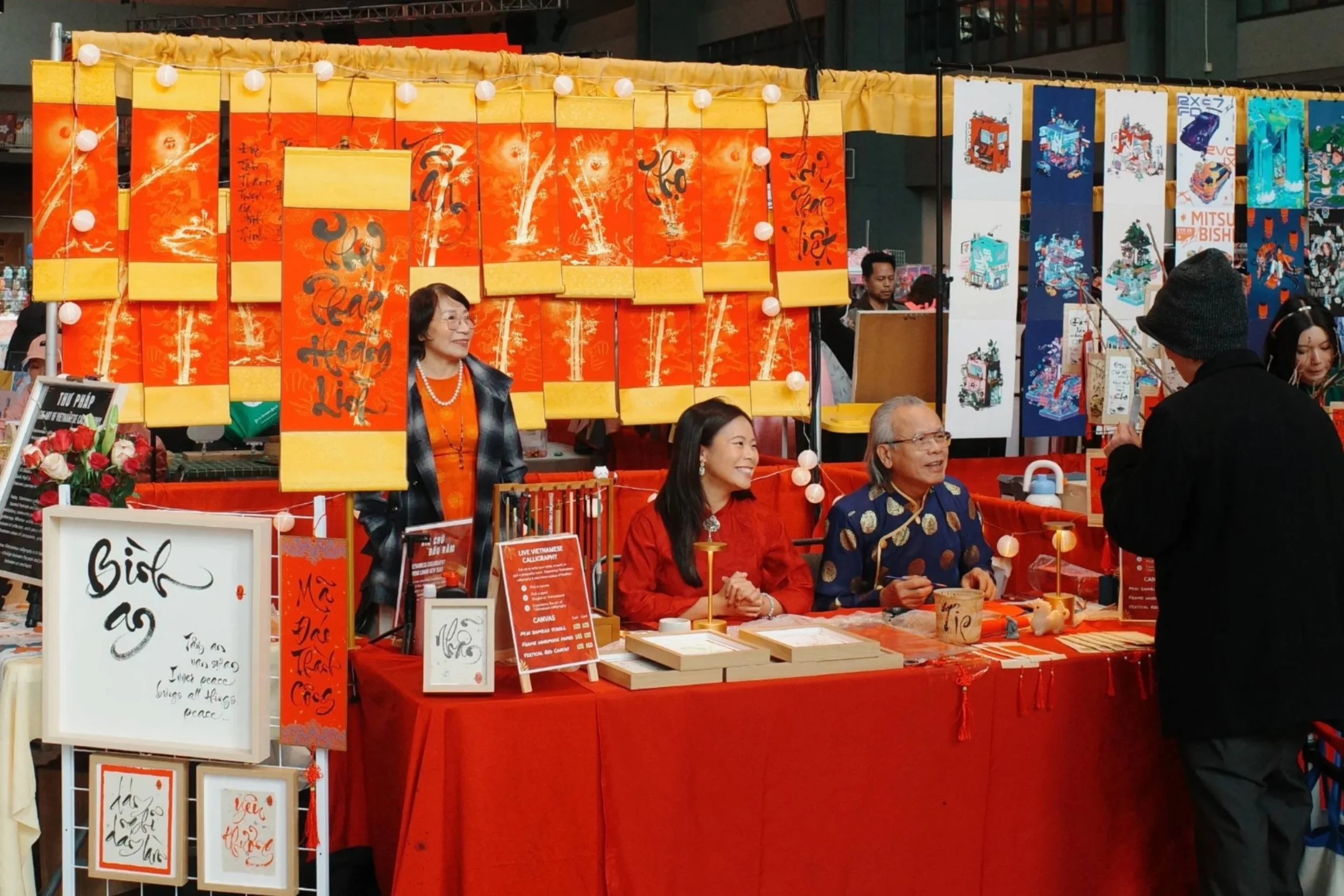 Linh Nguyen and her family at their Vietnamese calligraphy booth at Tết in Seattle 2025, surrounded by vibrant orange and gold calligraphy scrolls hanging in the background.
