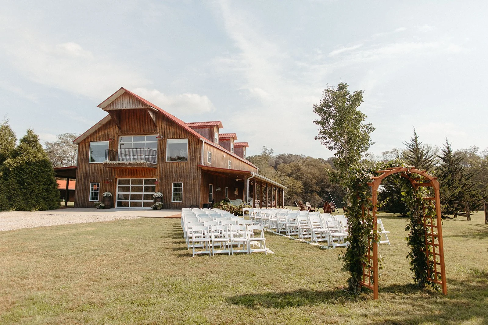 A rustic wooden barn converted for a wedding ceremony, with multiple rows of white chairs set up on the lawn, a floral arch at the entrance, and trees and open sky in the background.