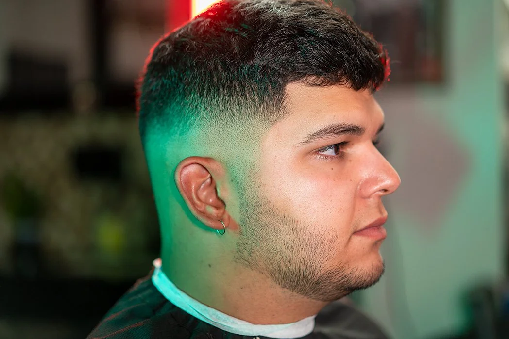 Profile of a young man with a high fade haircut, light beard, and a small earring, sitting in a barber shop with colorful lighting.