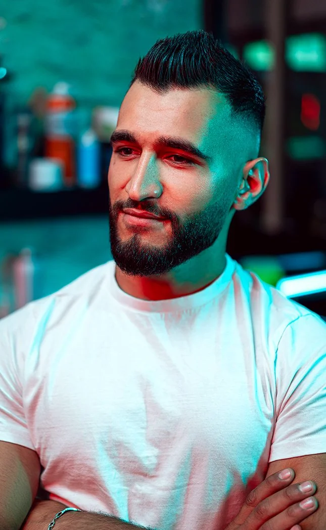 A man with a well-groomed beard and a modern hairstyle, wearing a white t-shirt, standing with arms crossed in a barbershop or salon with various bottles and tools in the background.