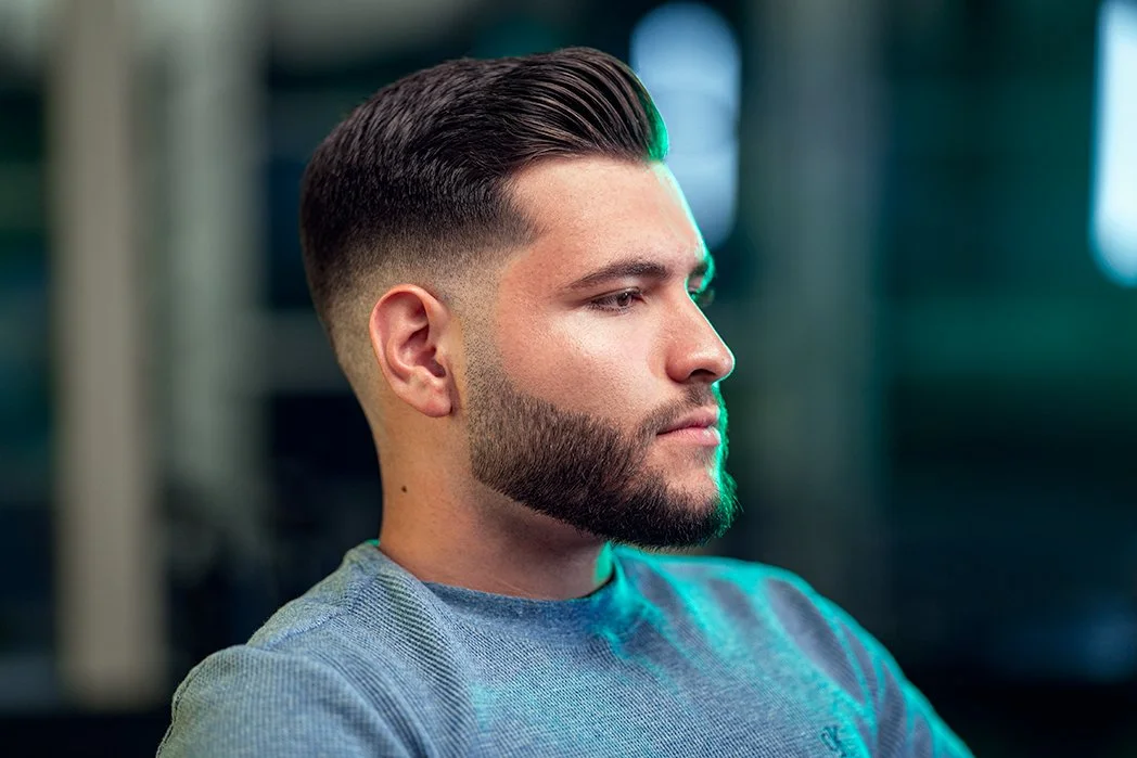 Side profile of a man with styled hair and a full beard, wearing a grey shirt, in an indoor setting with soft lighting.