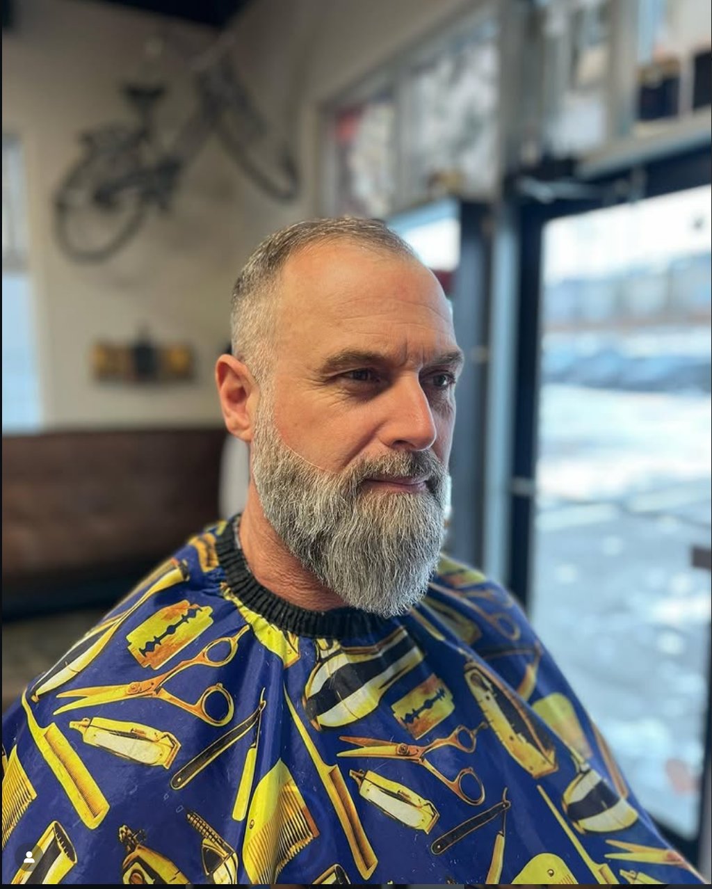 A middle-aged man with a beard and short hair sitting in a barbershop, wearing a cape with a hairdressing tool pattern, looking to the side near a window with a blurry outdoor scene in the background.