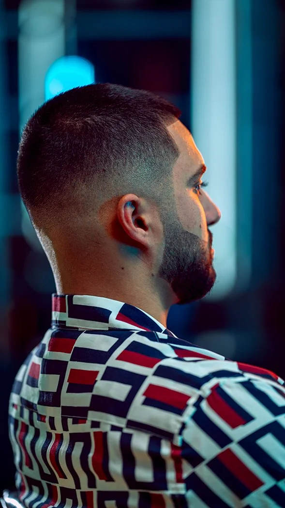 Side profile of a man with a buzz cut hairstyle and a well-groomed beard, wearing a patterned shirt with red, white, and navy geometric shapes, in a dimly lit environment with blue and teal lighting.