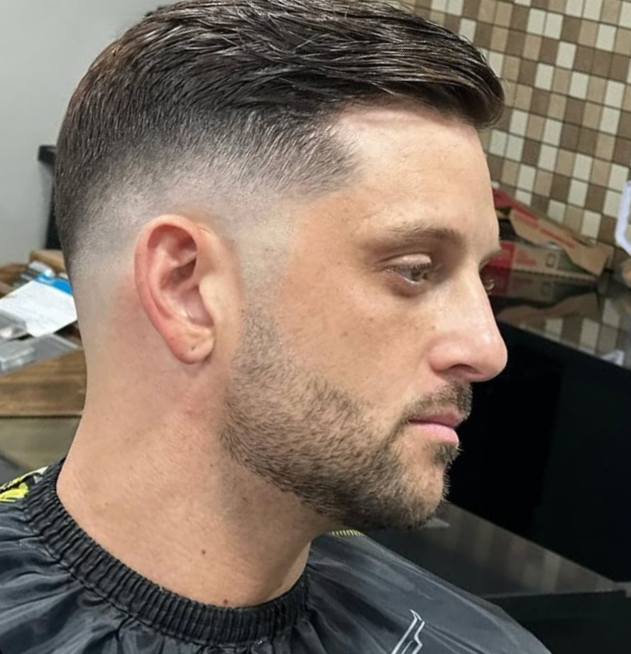 Side profile of a man with a fresh haircut, featuring a fade hairstyle and neatly trimmed beard, sitting in a barber shop.