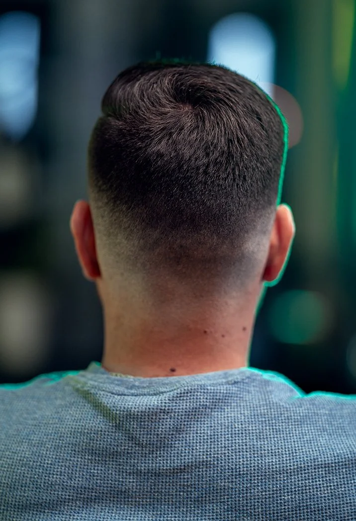 Back of a person's head with a short, neatly styled haircut, wearing a gray shirt, blurred background with green and blue hues.