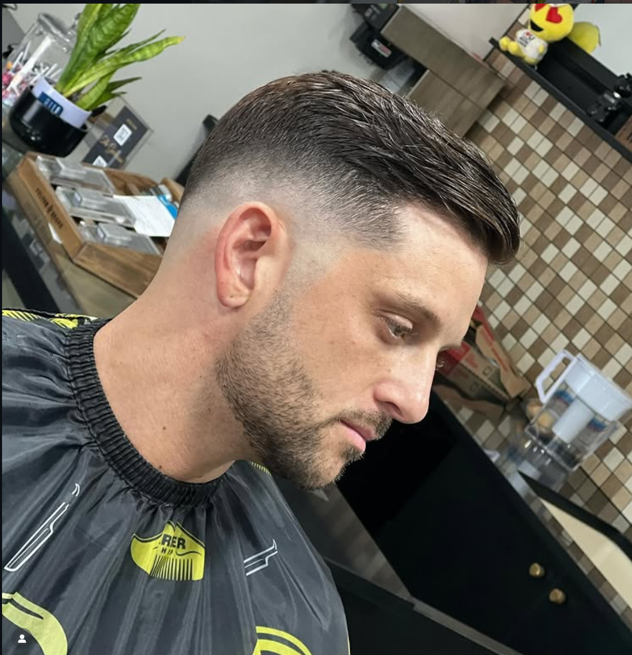 A man with a fresh, clean fade haircut and neatly combed hair, sitting in a barbershop chair wearing a black and yellow barber cape.