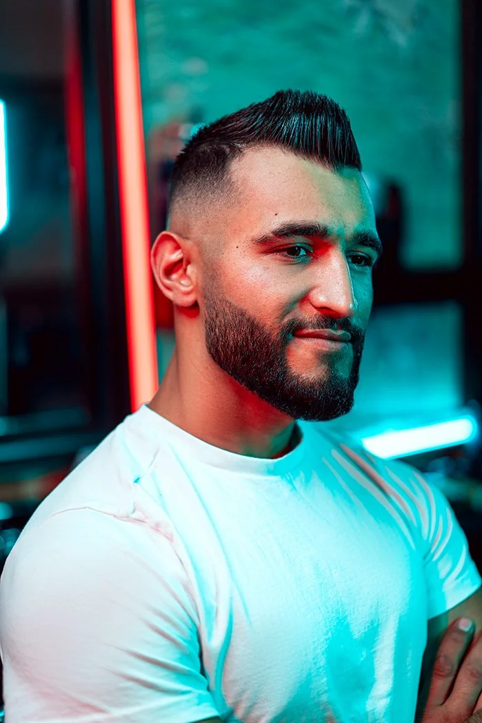 Young man with a beard and short hairstyle, wearing a white t-shirt, standing in a setting with colorful neon lighting.