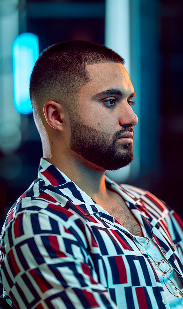 Profile of a young man with short dark hair and a beard, wearing a patterned shirt in a setting with blue lighting.