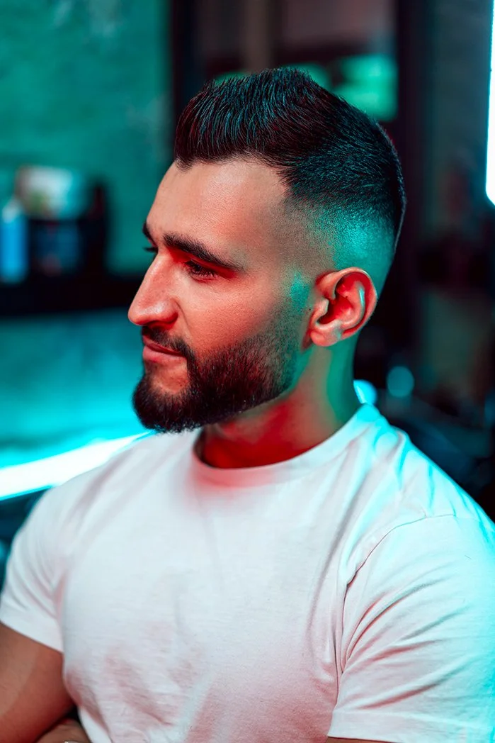 Side profile of a young man with a styled haircut and beard, wearing a white t-shirt, in a room with colorful lighting.