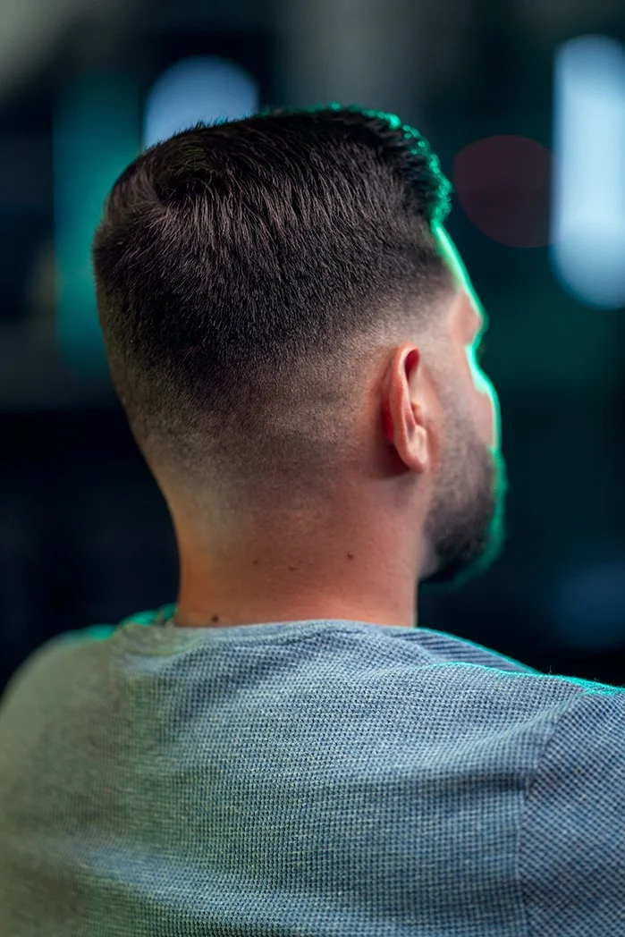 Back view of a man with a fade haircut, wearing a light grey shirt, in a dimly lit environment with blurred background lights.