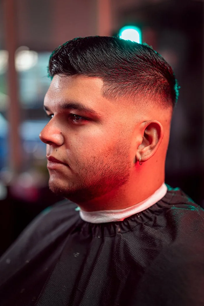 Young man with dark hair and a beard, wearing a black barber cape and a small hoop earring on his left ear, sitting in a barbershop with blurred background and neon lighting.