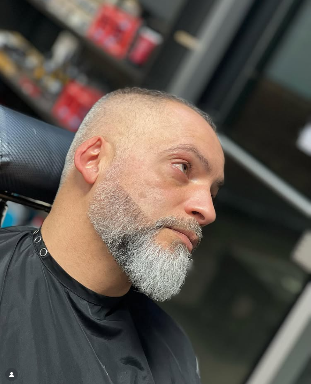 Man with a freshly shaved head and a grey beard, sitting in a barber's chair in a barbershop.