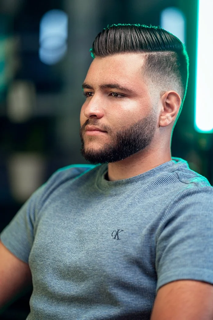 A young man with styled dark hair and a full beard, wearing a gray T-shirt, sitting indoors with blurred background.