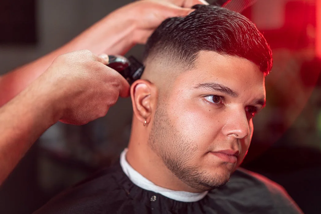 A man getting a haircut at a barbershop, with a barber trimming the sides of his hair with clippers, while the man looks forward in a black barber cape.