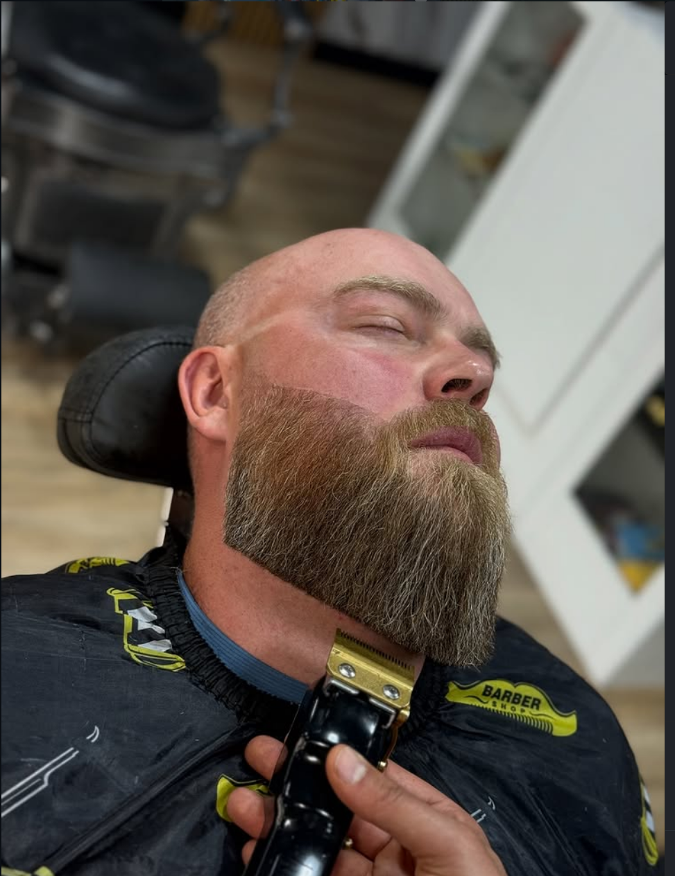 A man with a beard and closed eyes is lying back in a barber chair, getting a shave or close trim with an electric clipper.