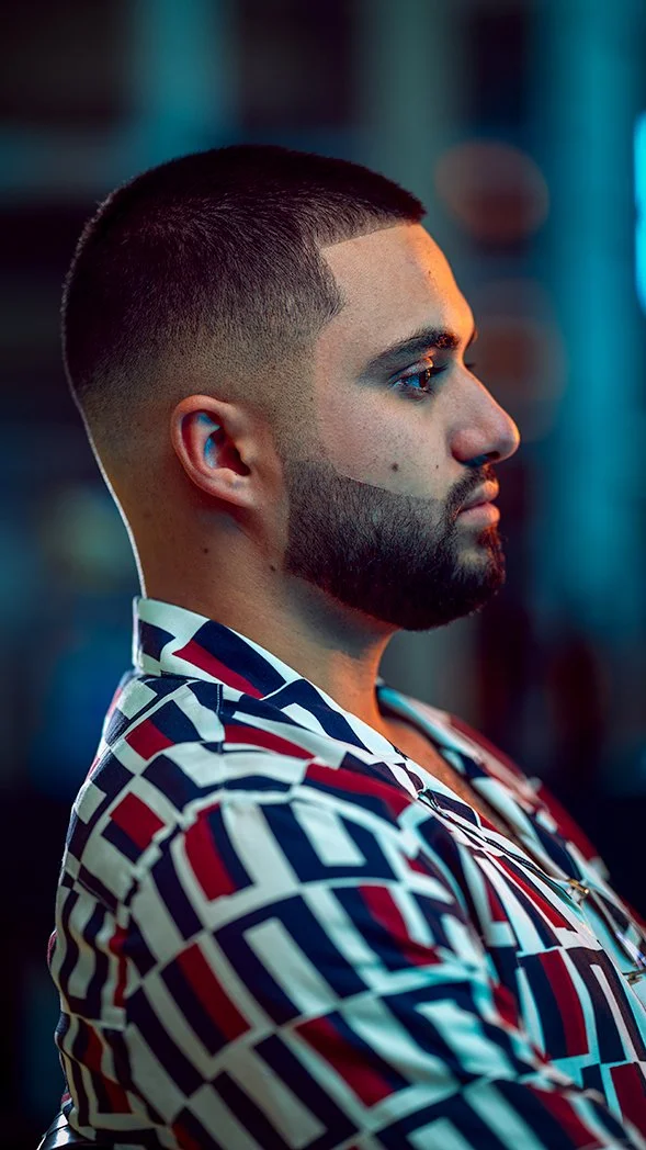 A man with a short haircut and full beard, wearing a patterned shirt, is shown in profile against a blurred colorful background.
