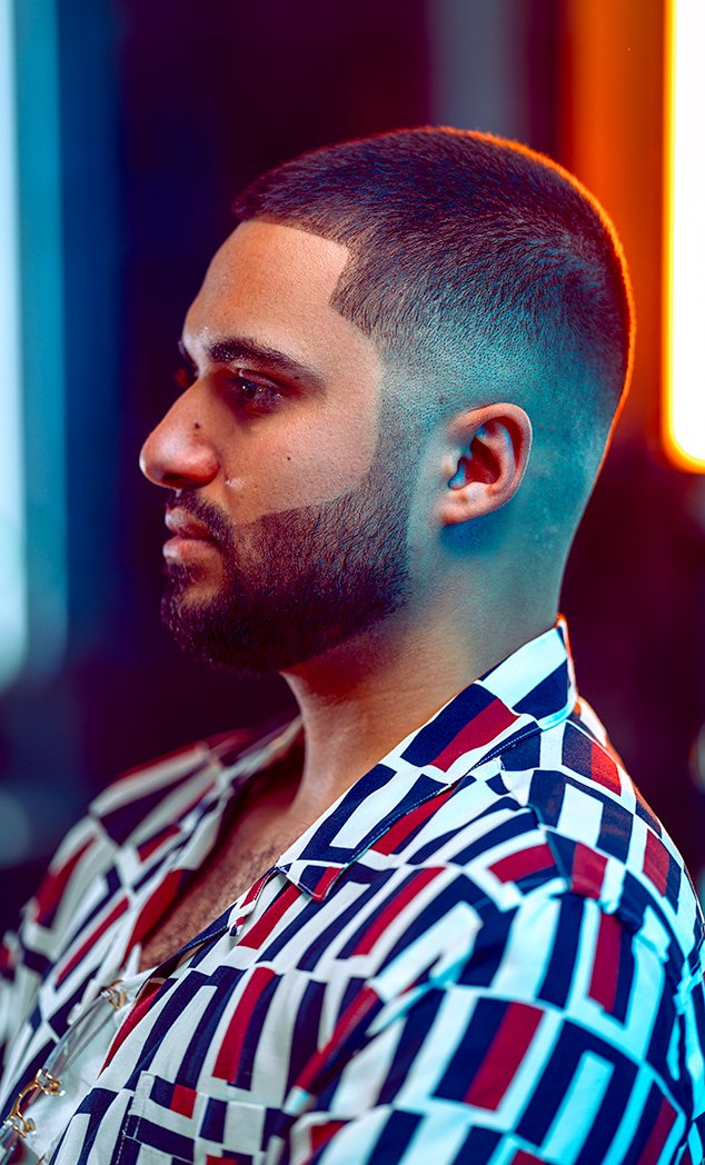 Profile of a man with a neatly groomed beard and short haircut, wearing a patterned shirt, in a setting with colorful lighting.