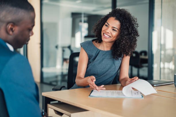 A woman with curly hair smiling and talking to a man in an office setting, with documents on the table.