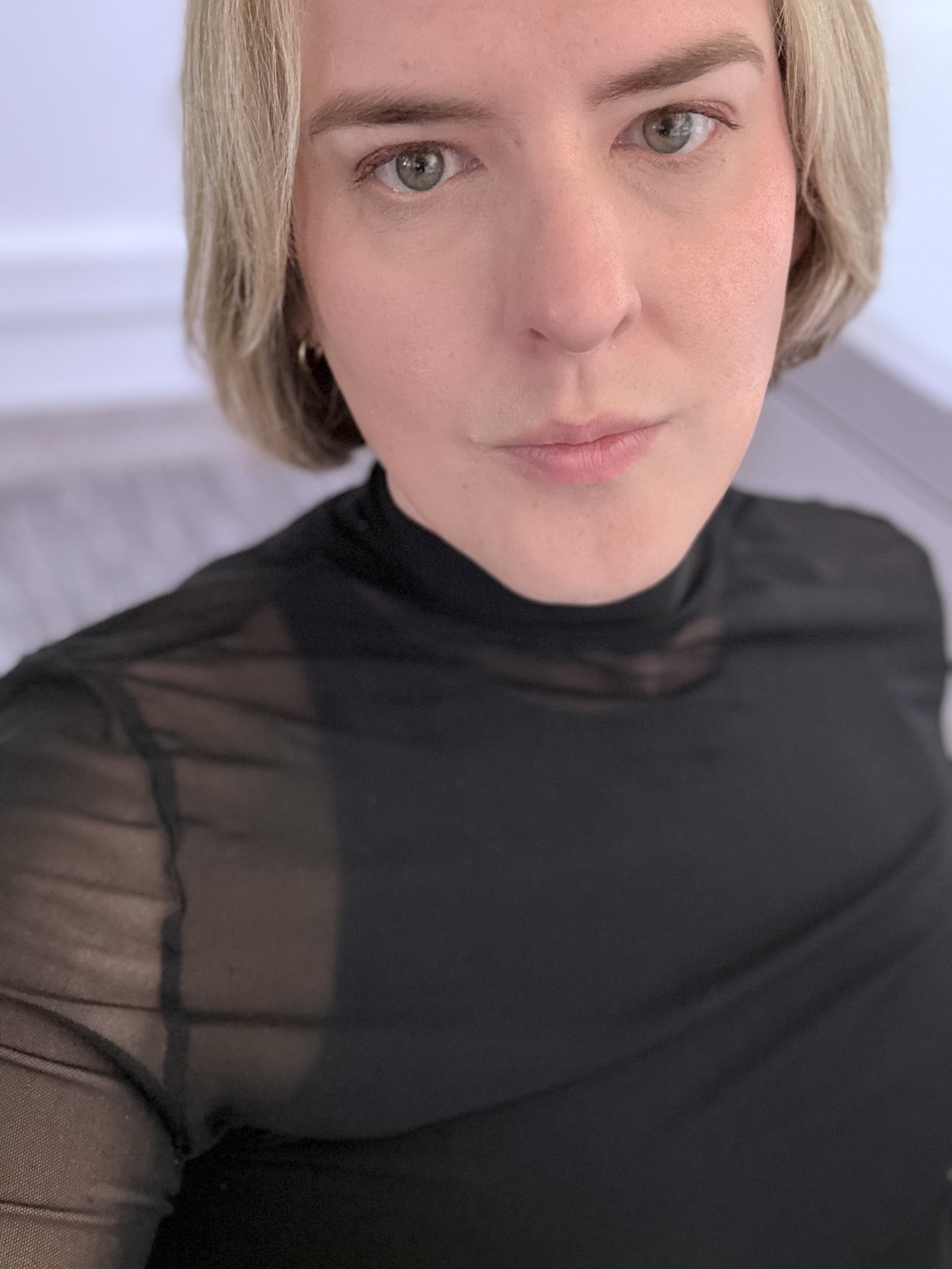Soft portrait of Pauly with a blonde bob and sheer black top, photographed in gentle natural light with a calm, intimate expression that evokes elegance and quiet strength.