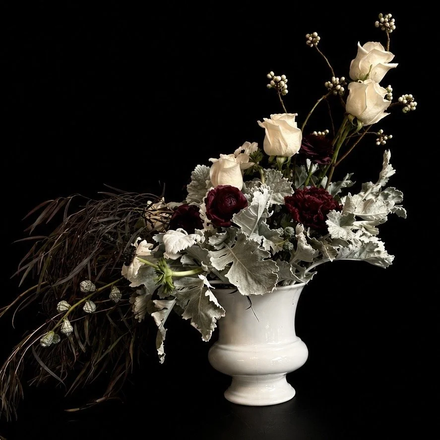 Avant-garde floral arrangement in a white ceramic vase with ivory roses, deep burgundy blooms, silver foliage, and cascading dark grasses against a black background.