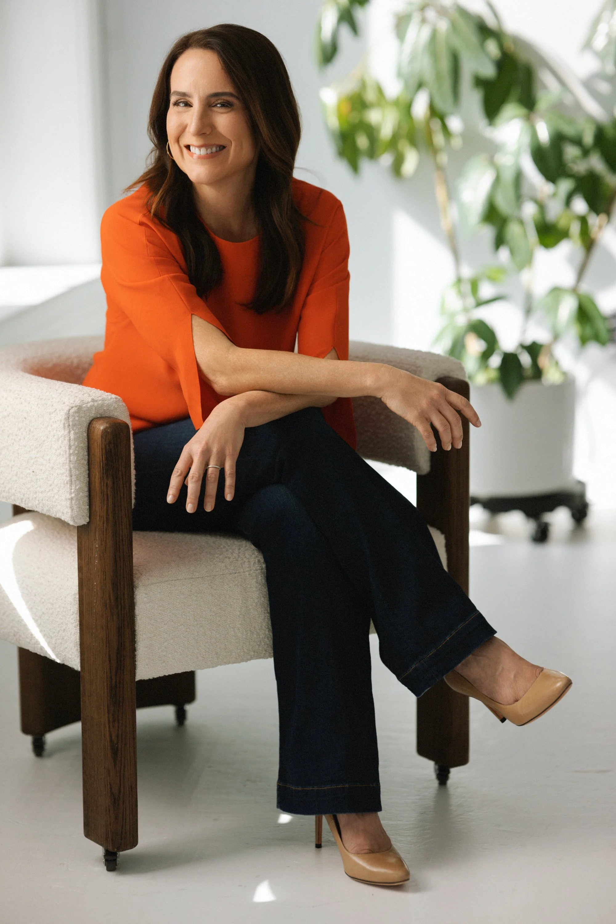 Journalist Janet Adamy sitting on a beige armchair, smiling at the camera, wearing an orange top, dark jeans, and nude heels, with a potted plant in the background.