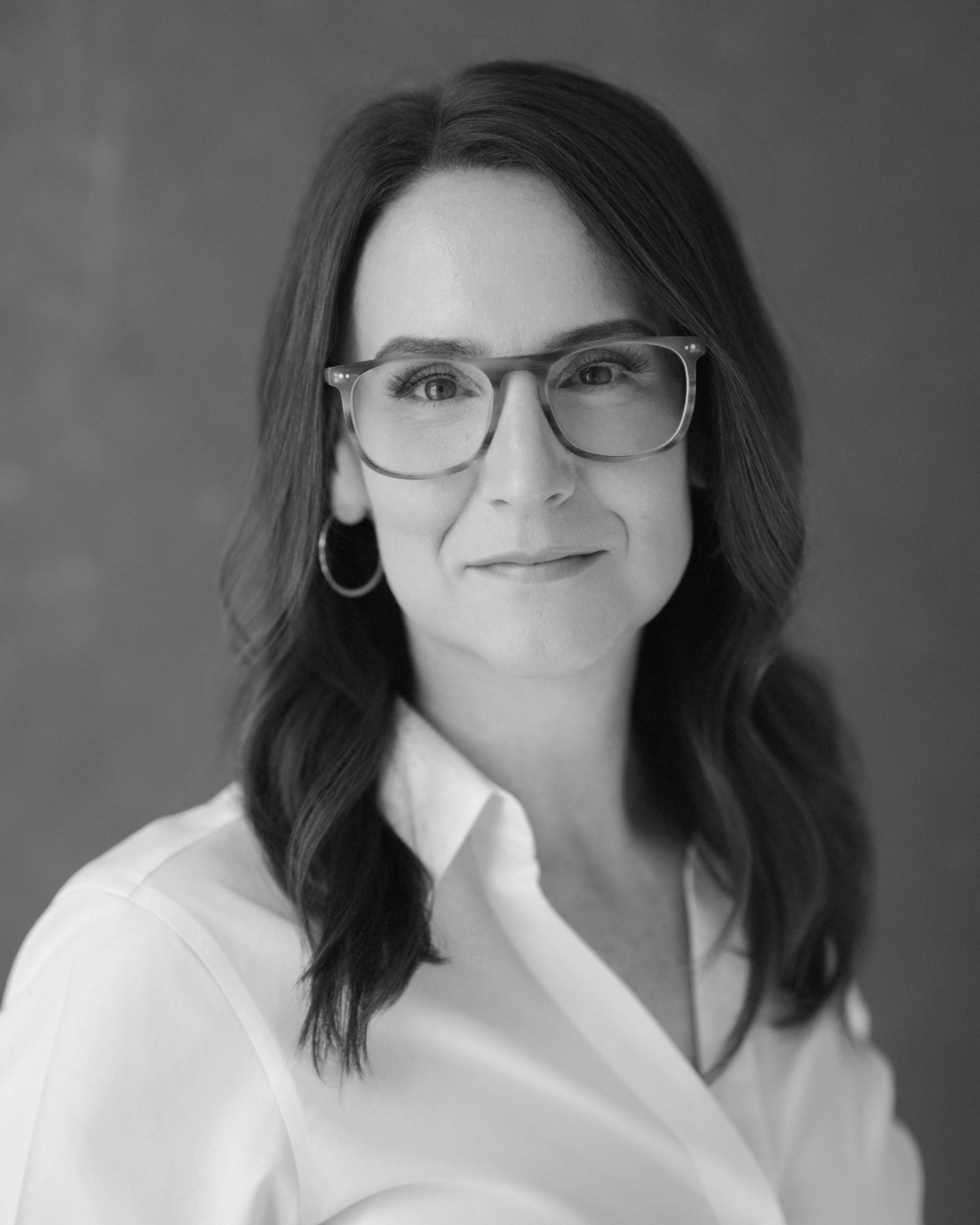Black-and-white portrait of journalist Janet Adamy, smiling gently, with wavy dark hair, glasses, hoop earrings, and a white blouse.