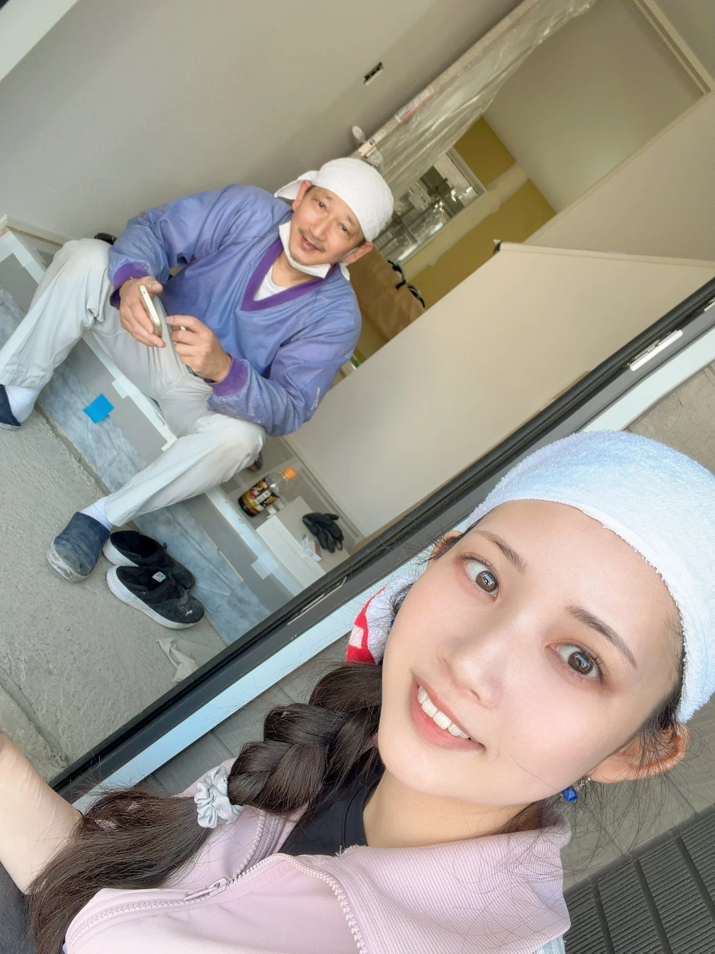 二人の女性がバスの外側の鏡越しに自撮りをしている。女性の一人は笑顔で若々しい顔立ち、もう一人は工事作業員の男性と交流している。