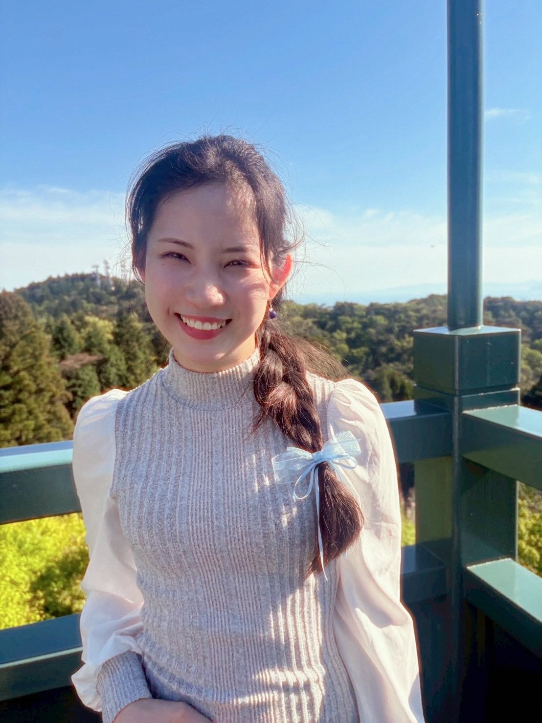 青空の下で微笑む女性の写真。背景には緑の森林と遠くの海が見える、展望台やバルコニーから撮影された風景写真です。