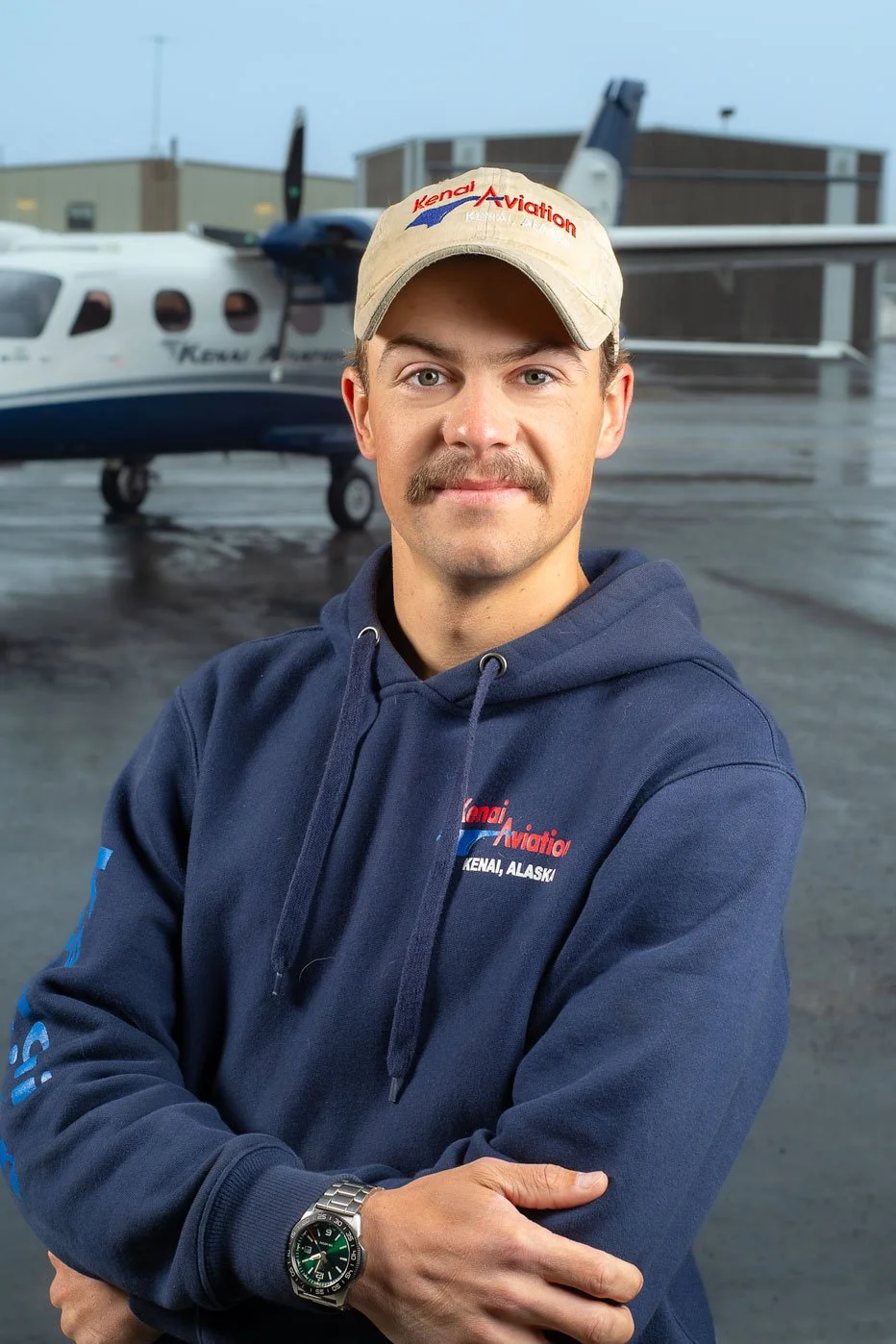 Man wearing a beige Kenai Aviation cap and navy hoodie standing in front of a small airplane on an airport tarmac.