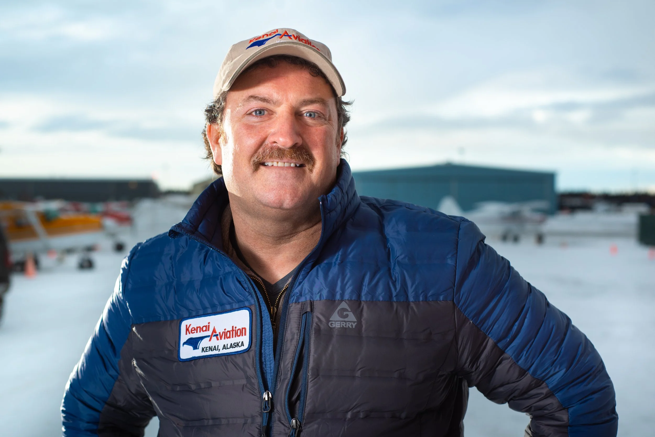 A man in outdoor winter clothing standing outside in a snowy area with planes and a large hangar in the background.