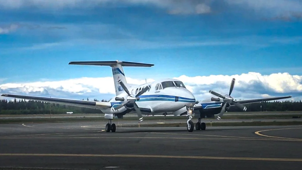 A white and blue twin-engine propeller airplane on the runway with a cloudy sky and trees in the background.