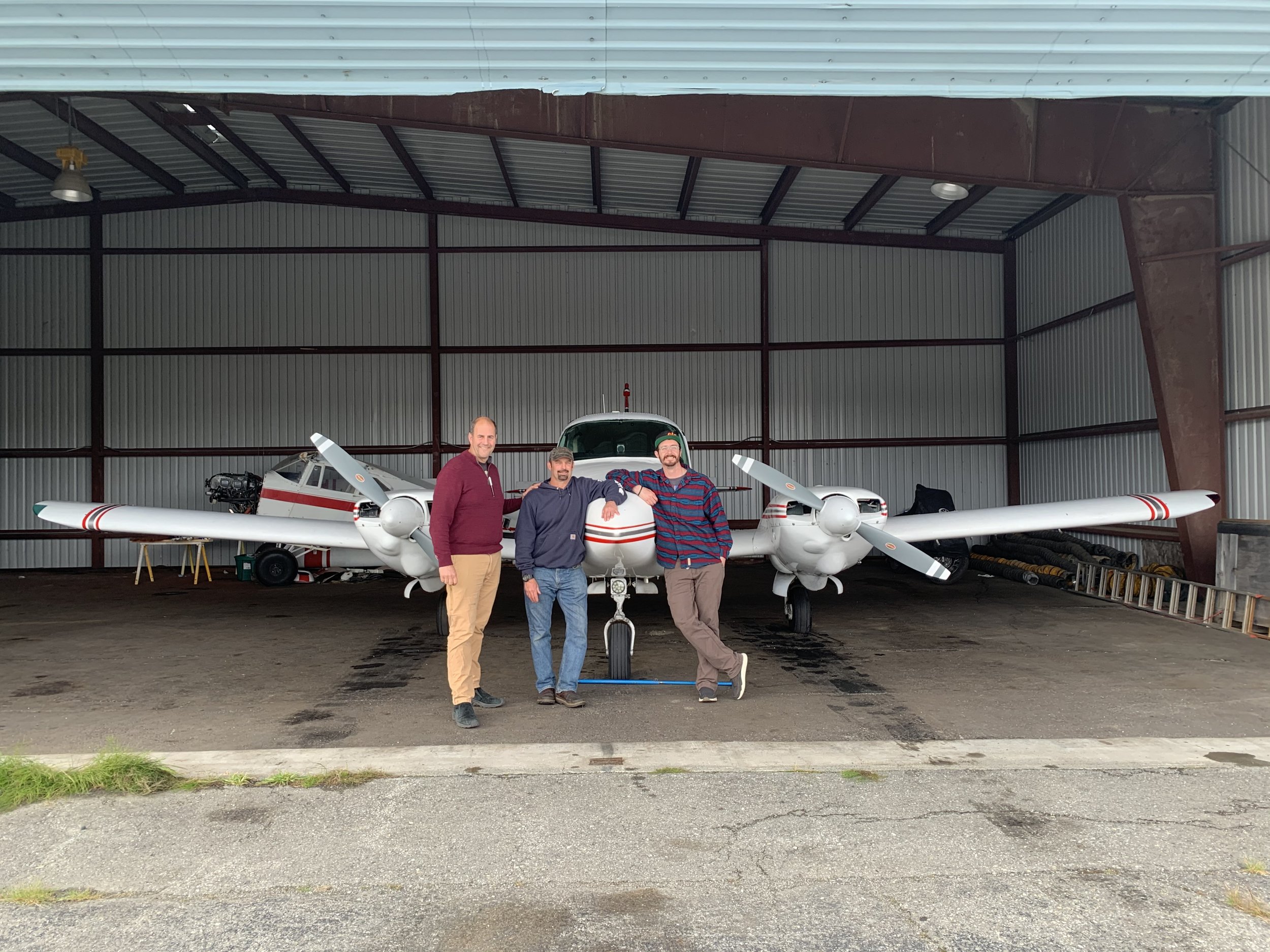 Three men standing in front of a twin-propeller airplane inside a hangar.