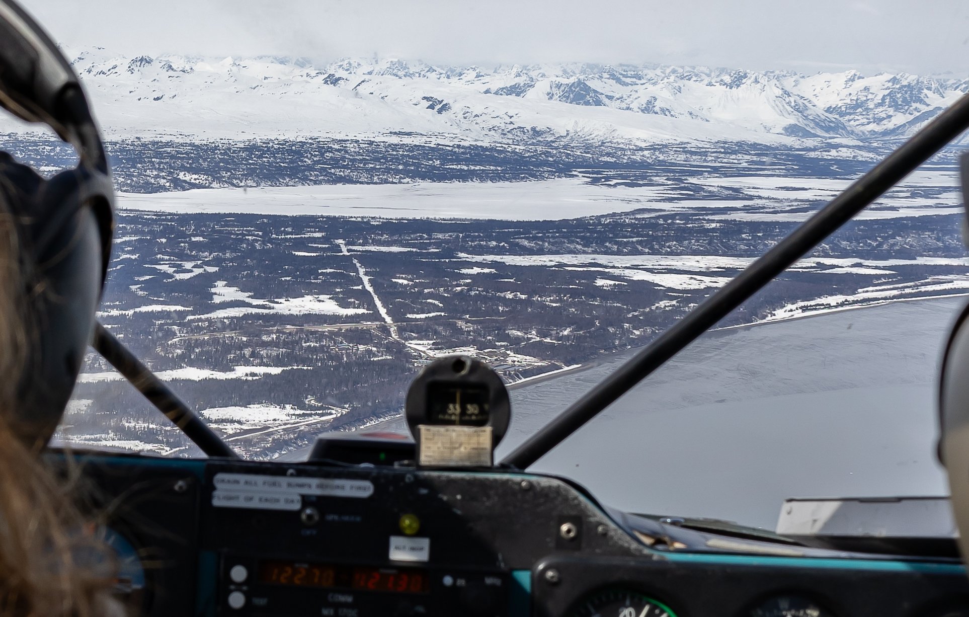 View from inside a helicopter cockpit showing snow-covered landscape and mountains below during winter.