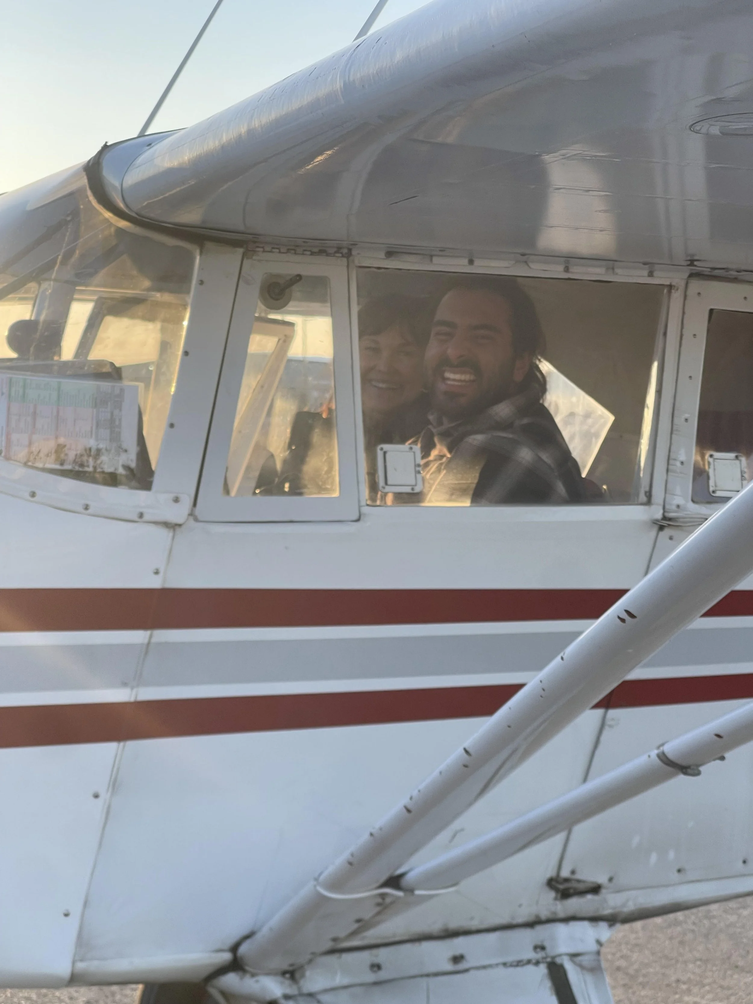 Three smiling people inside a small white airplane with red stripes, seen through the cockpit windows during sunset.