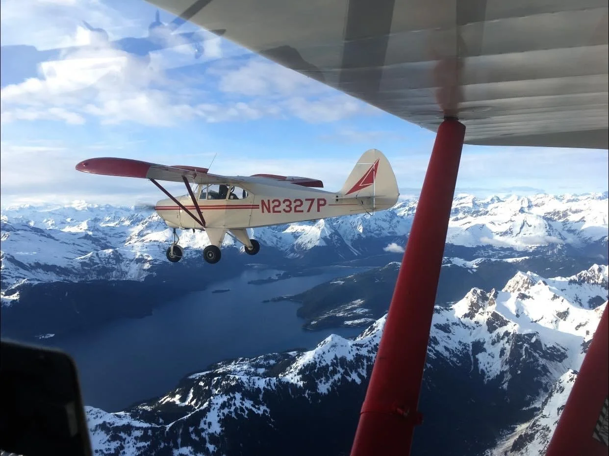 Small single-engine airplane with registration N2327P flying over snow-covered mountains and a fjord, viewed from another aircraft.