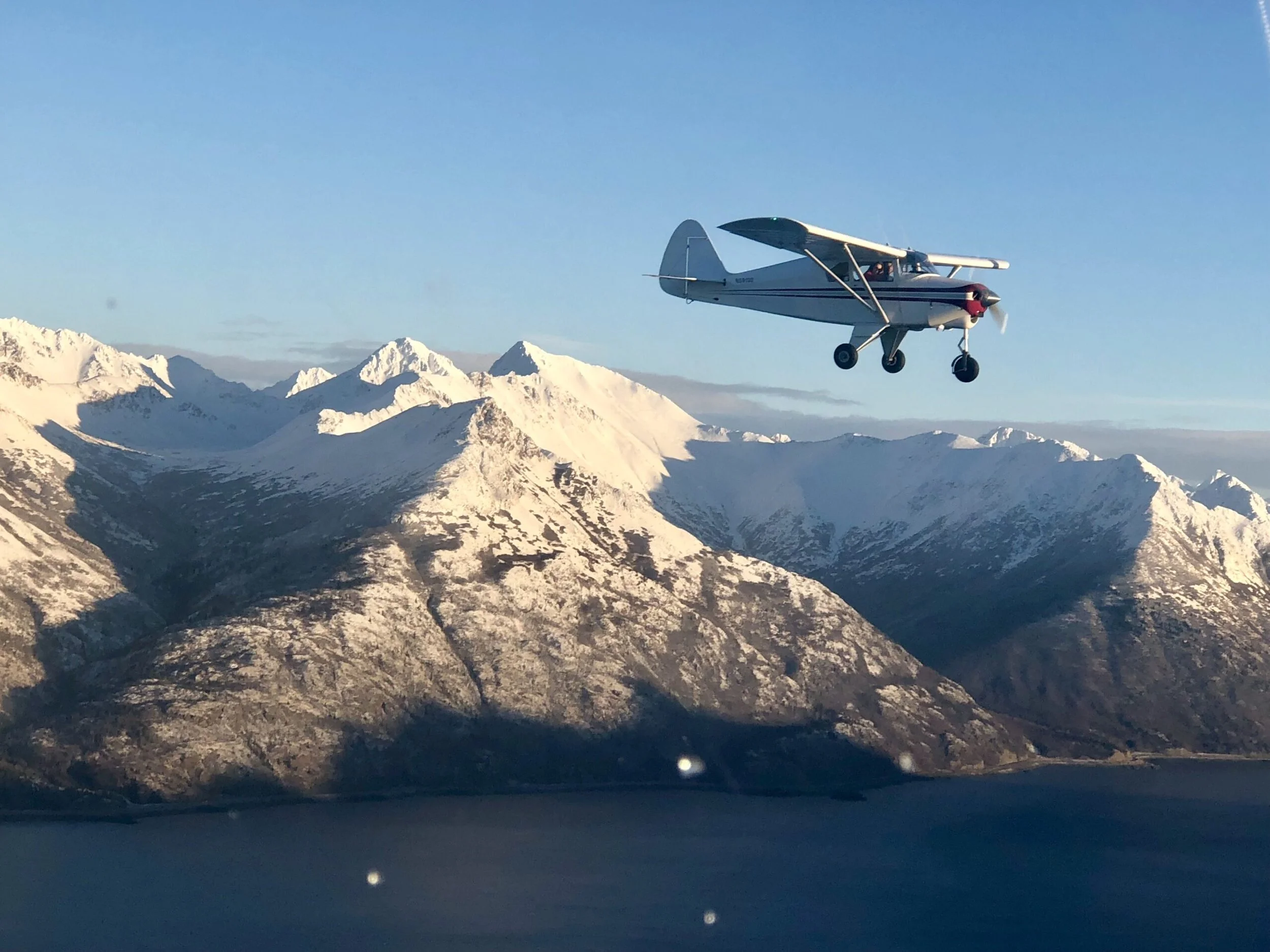 Small airplane flying over snow-covered mountains and a body of water under a clear blue sky.