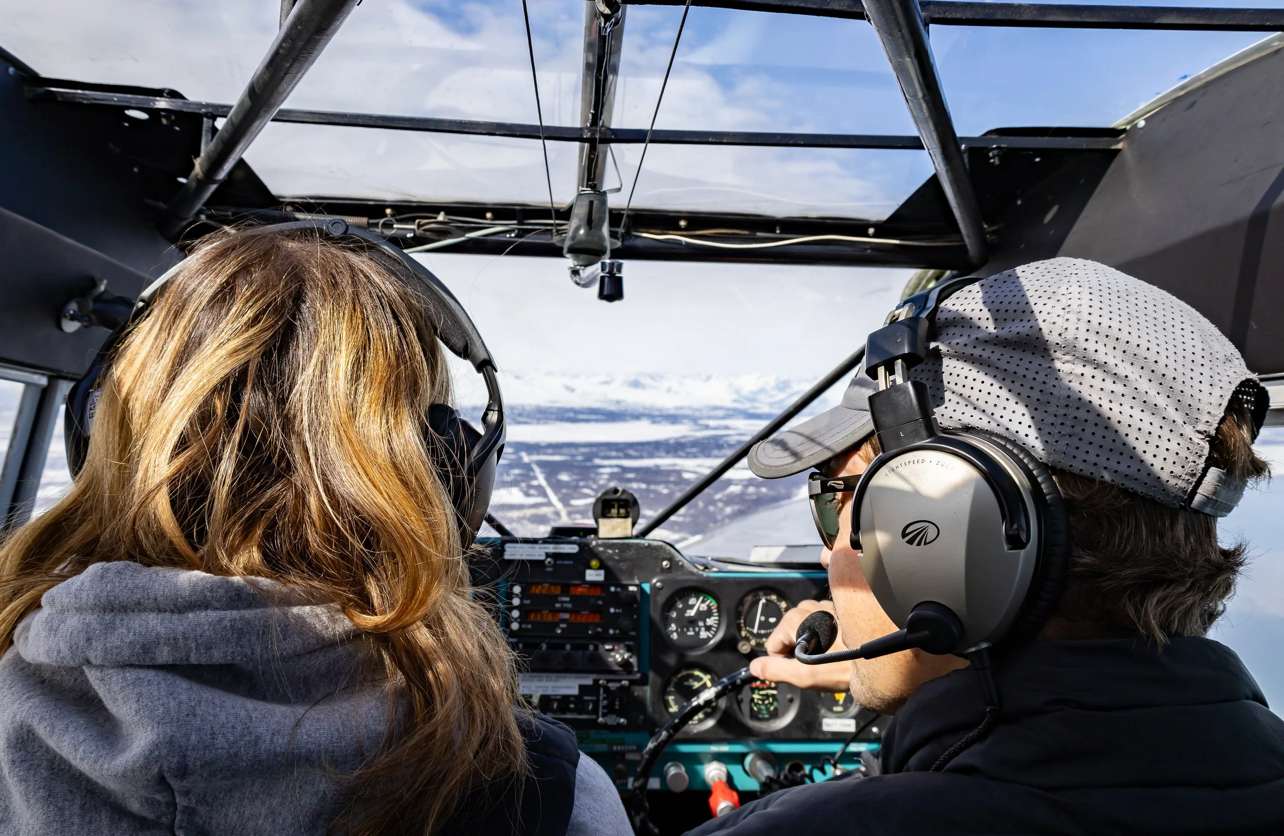 Two people inside an airplane cockpit flying over snowy mountains, both wearing headsets and sunglasses.